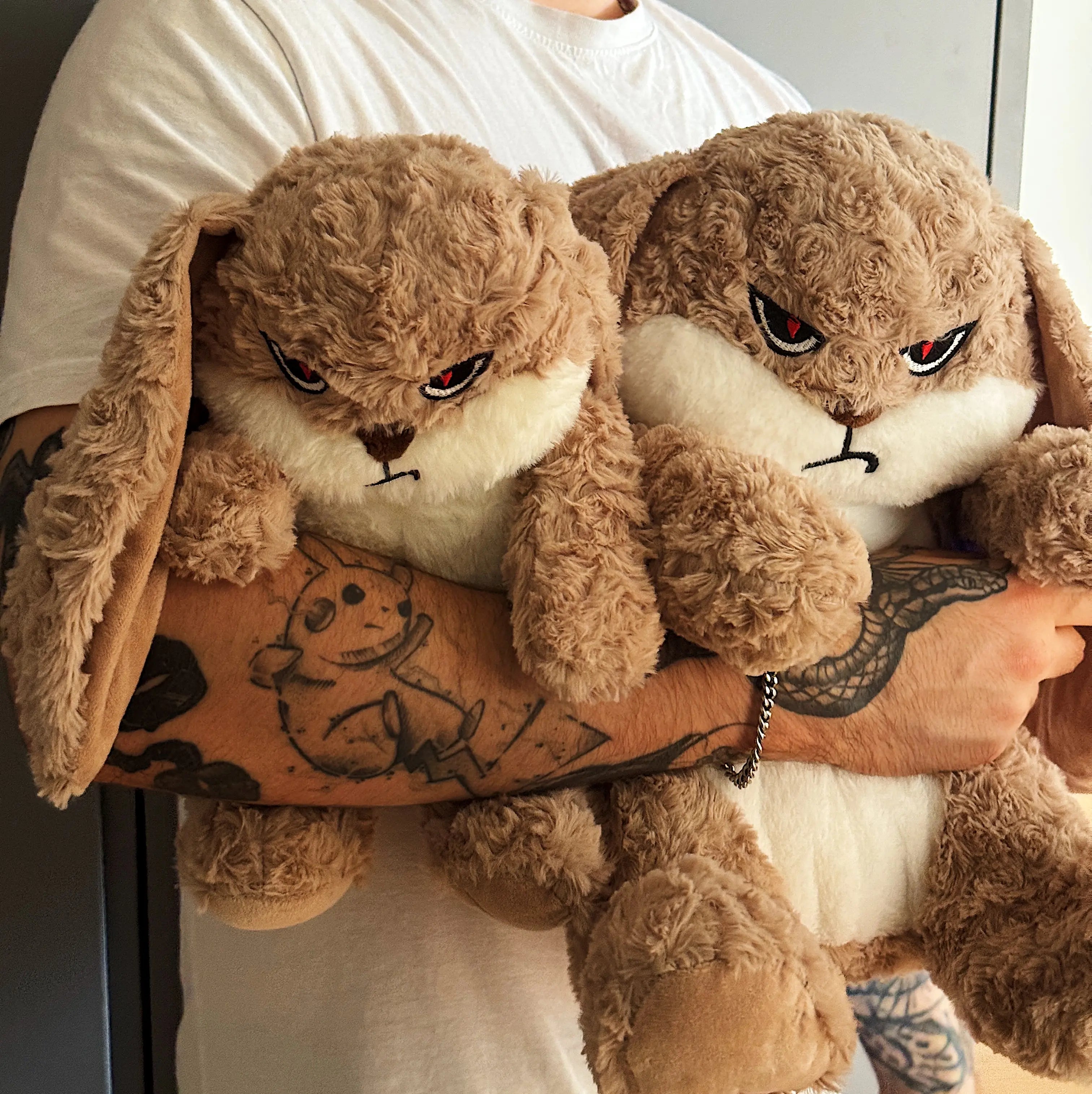 Two plush toys resembling angry bunnies held by a person with tattoos.