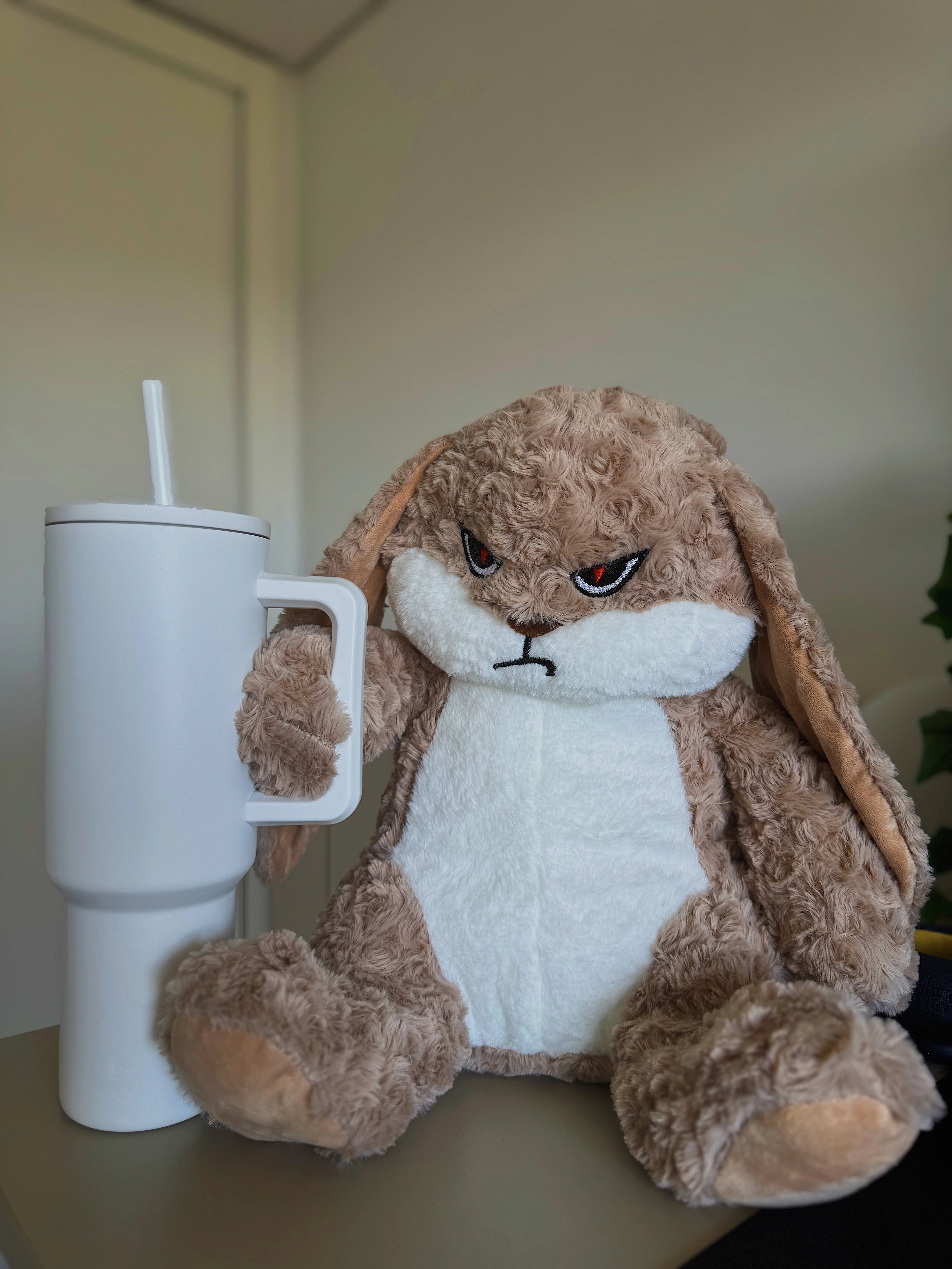 Plush bunny holding a white tumbler with a straw on a neutral background